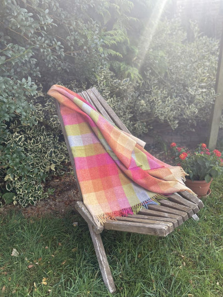 A multicolor plaid throw blanket with fringe is draped over a weathered wooden garden chair on a grassy lawn, with green bushes in the background.