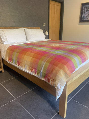 A bed in a bedroom setting with a colorful plaid throw blanket draped over it, white pillows, a light wood bed frame, dark gray tiled flooring, and a gray wall behind.